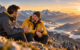 Zwei Männer im Gespräch auf einem felsigen Berg mit Blick auf die Alpen im Abendlicht. Einer trägt eine gelbe Jacke.